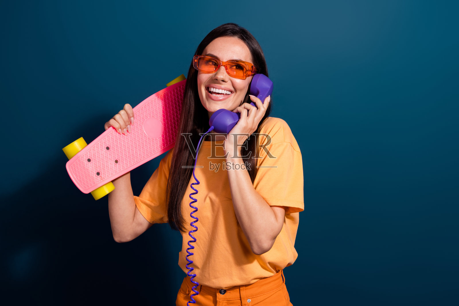 时髦女人的肖像穿着橙色t恤打电话旧电话邀请朋友骑滑板孤立在深蓝色的背景照片摄影图片