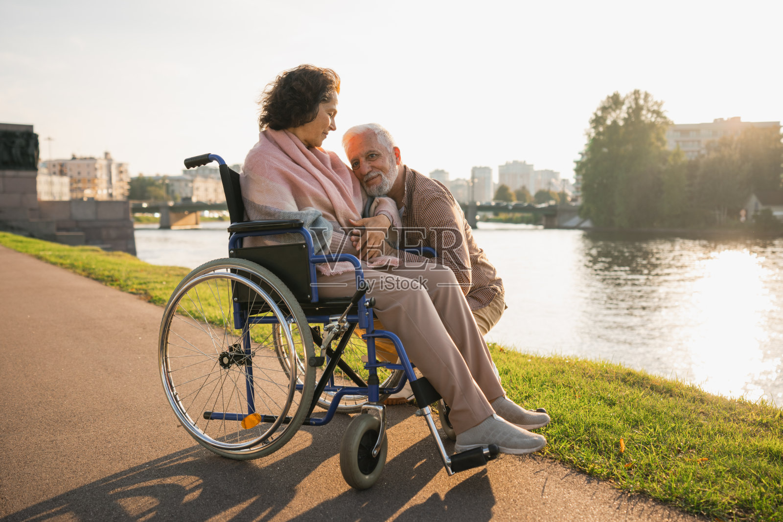 坐在轮椅上的老年妇女与照顾者老人在路上行走。老年家庭夫妇在户外为残疾人士提供的椅子上照顾瘫痪妇女。康复。照片摄影图片