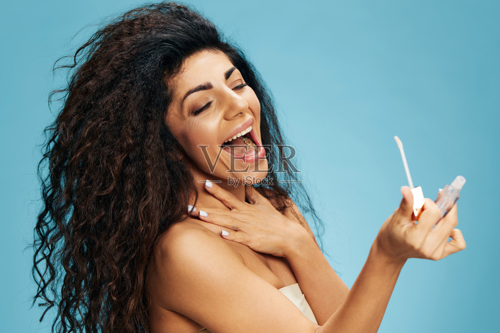 惊讶快乐迷人卷曲的拉丁女士看唇彩口红的姿态孤立在柔和的蓝色背景。彩妆唇部产品广告自然美妆概念工作室肖像复制空间照片摄影图片