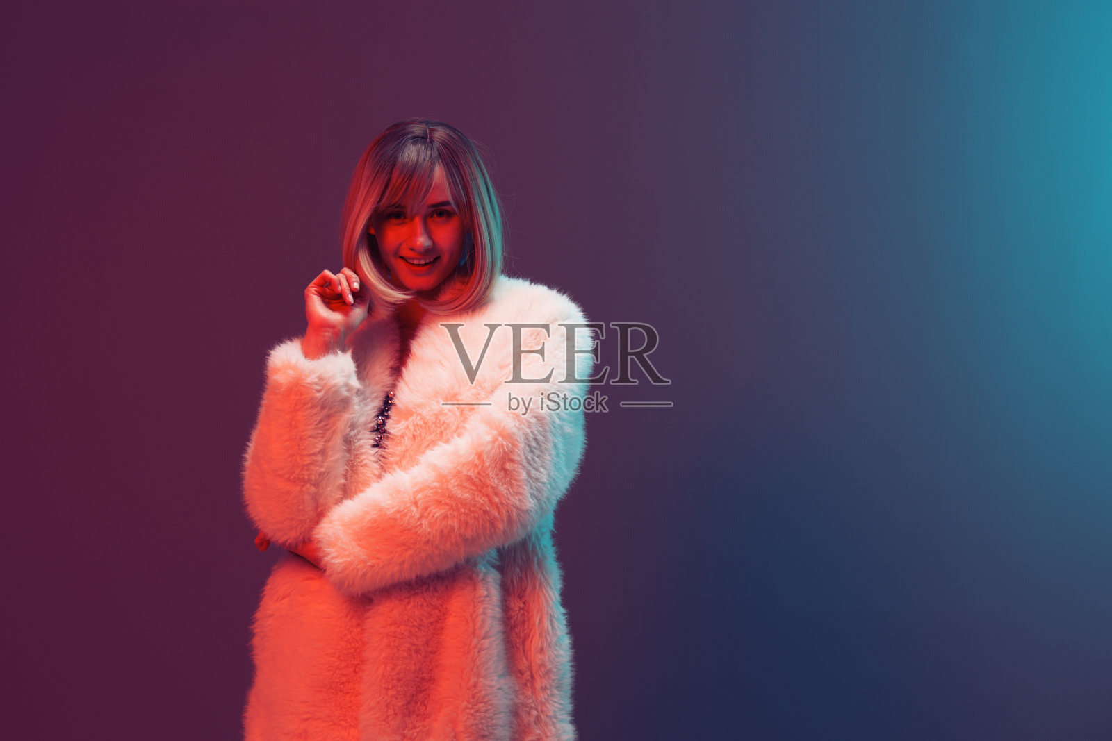 可爱可爱的金发女郎，穿着蓬松的毛皮大衣，闪闪发光的裙子，在镜头前调情微笑，在蓝色绿松石红色的光工作室背景下摆姿势。霓虹灯派对时尚概念。复制空间横幅照片摄影图片