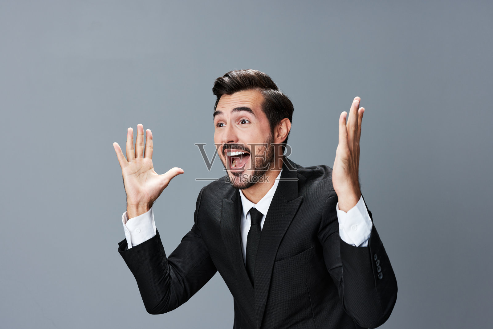 商人开心地笑着，咧着嘴，举起手，惊讶地在灰色的背景上穿着西装。照片摄影图片