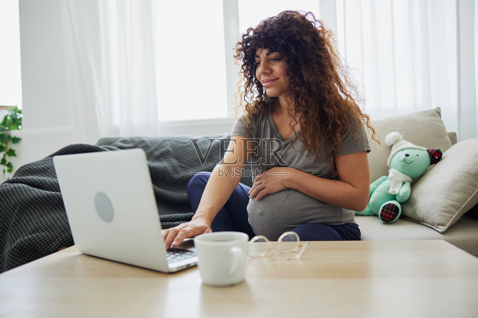 孕妇坐在家里的沙发上用笔记本电脑视频聊天，博主，自由职业者在网上工作。家庭服装，孕妇的生活方式，为分娩做准备照片摄影图片