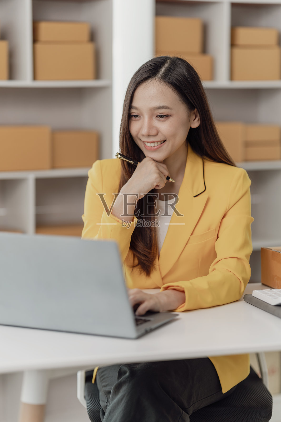 一名穿着围裙的亚洲自由职业者用笔记本电脑和盒子接收和检查网上订单，准备打包卖给客户。中小企业网上经营理念，垂直形象照片摄影图片