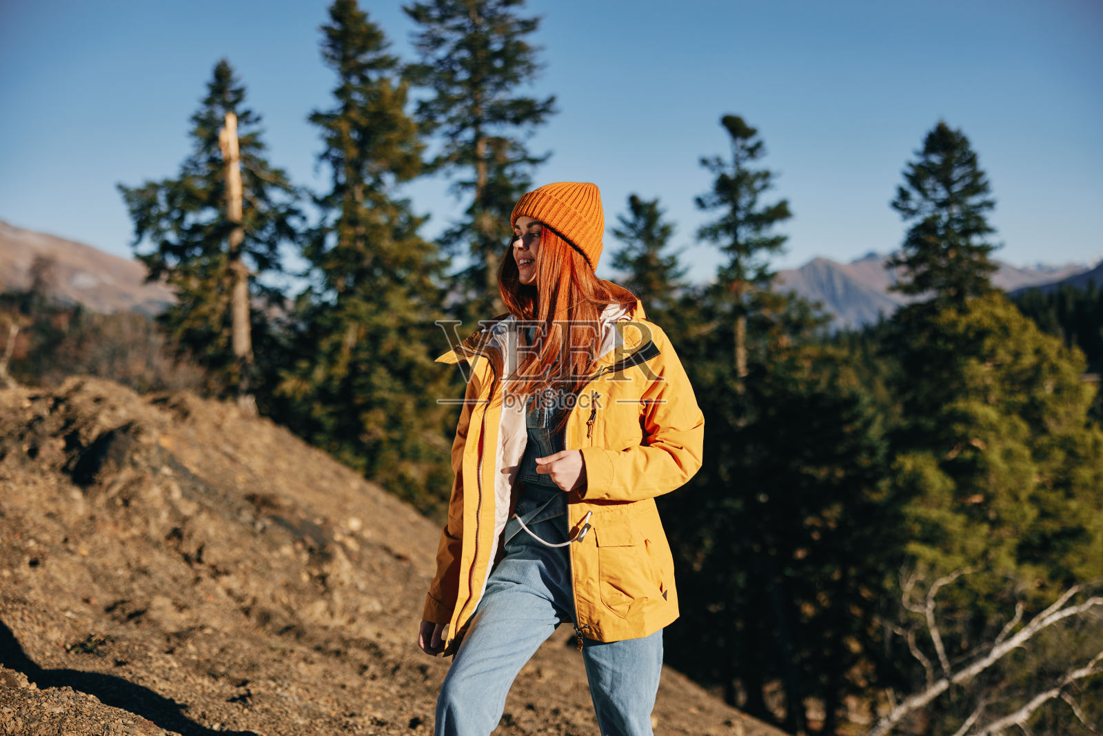 女人在秋日的山间徒步，披着黄披风，红发披风，站在树林和山前，在夕阳的余晖中微笑和幸福照片摄影图片