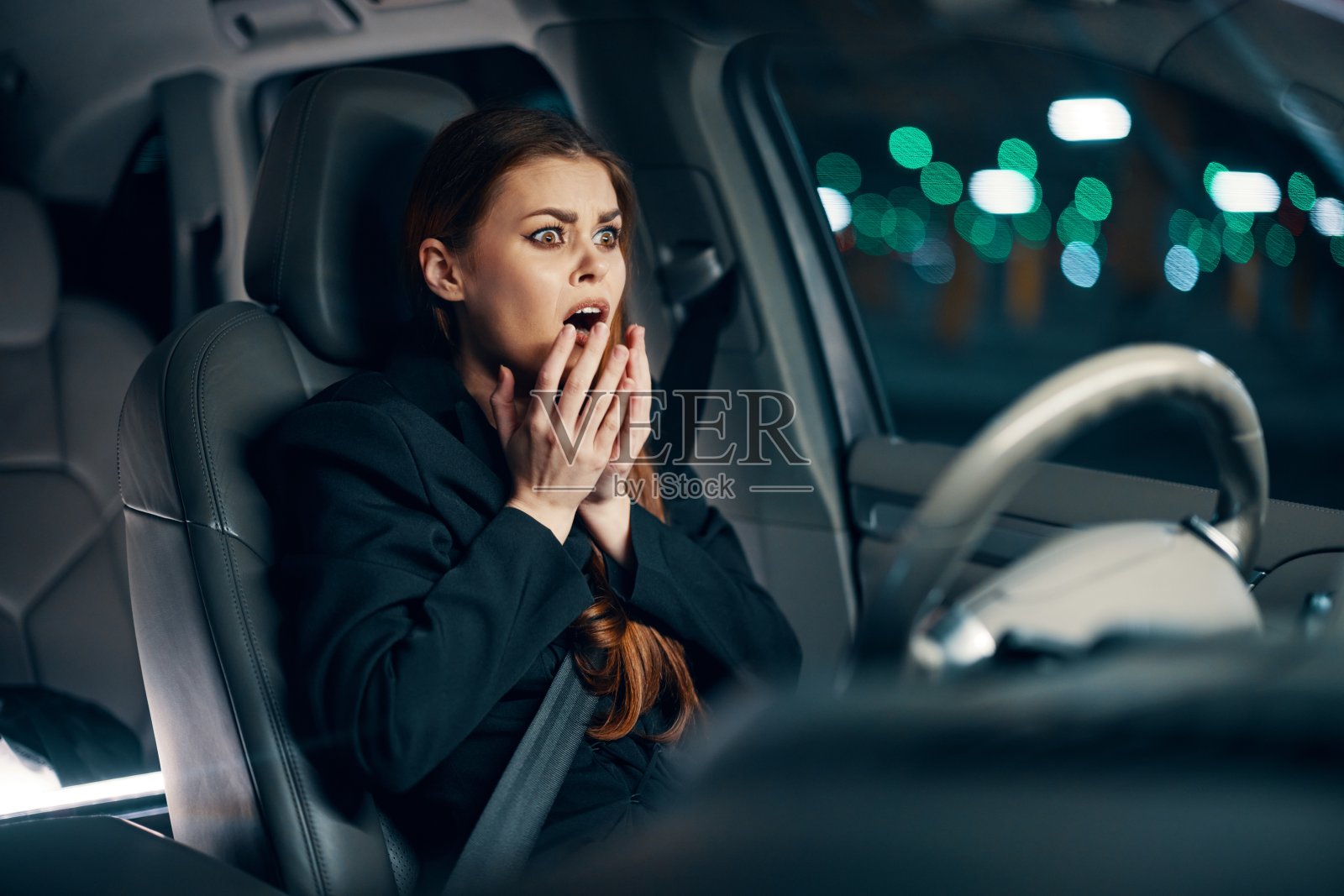 一位穿着黑色衬衫、系着安全带、用手捂着脸、情绪激动的女性坐在汽车的方向盘后面。夜间摄影照片摄影图片