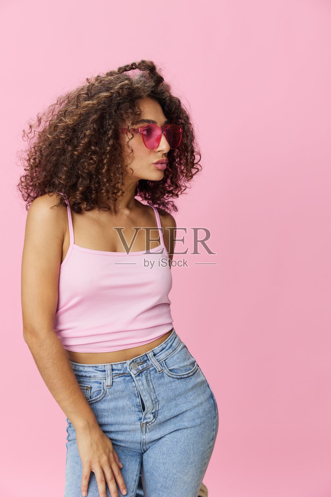 快乐的女人卷发在粉红色的背景上跳舞，夏天的粉红色t恤牛仔裤和眼镜，夏天的氛围，复制空间照片摄影图片