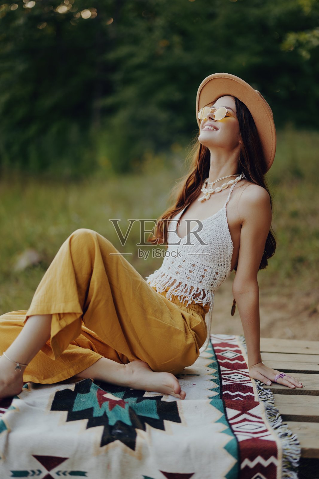 一个快乐的女孩，穿着环保服装，坐在湖边的格子草地上，戴着一顶嬉皮士式的帽子，惊奇地微笑着。秋天的生活方式照片摄影图片