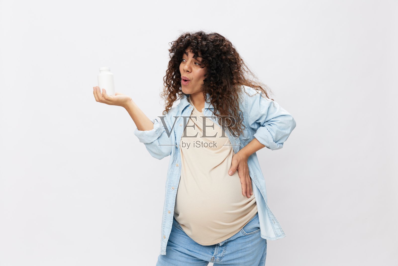 一位孕妇笑着露出牙齿，手里拿着一罐孕期维生素药片，α硫辛酸，维生素复合物在白色孤立的背景上照片摄影图片