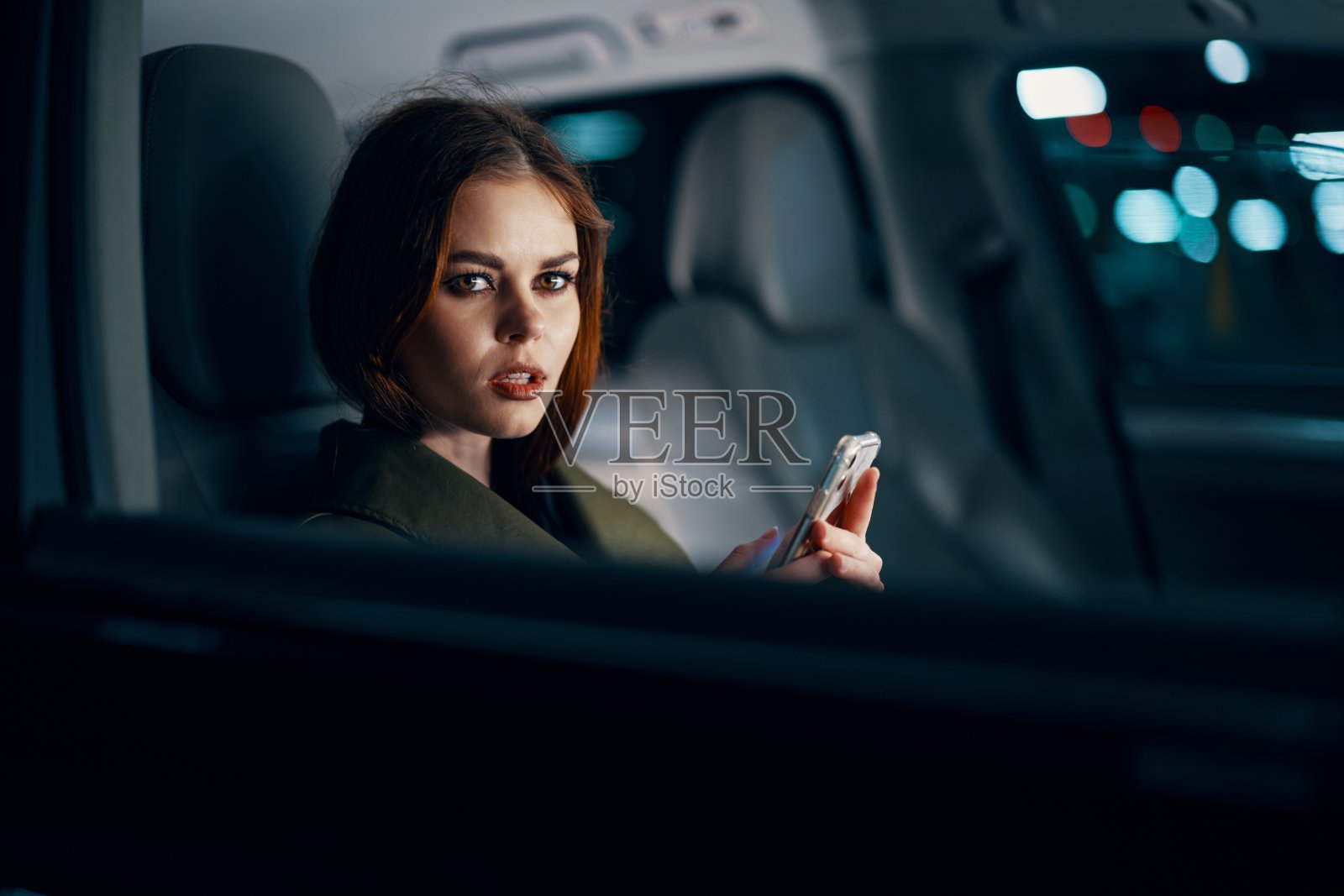 这是一幅近距离的横向肖像，一位穿着皮衣的时尚、奢华的女性晚上坐在一辆黑色汽车的副驾驶座位上，手里拿着智能手机，愉快地看着相机照片摄影图片