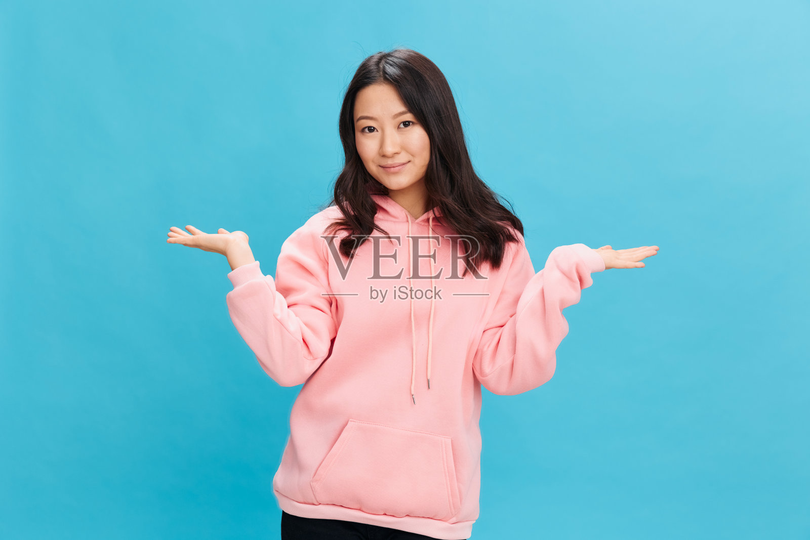 快乐可爱的亚洲学生年轻女士，穿着粉红色的连帽衫，在蓝色的工作室背景上展开双手，摆出孤立的姿势。最好的广告报价。人的日常情感概念照片摄影图片