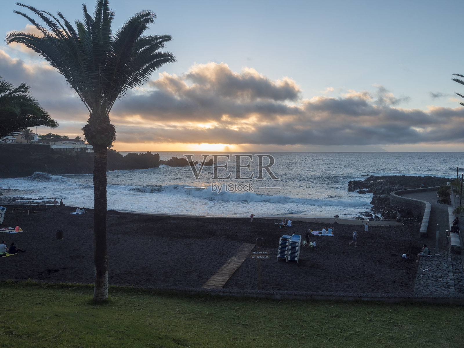 日落的黑色沙滩普拉亚德拉竞技场与戏剧性的橙色云和放松的游客和棕榈树。照片摄影图片