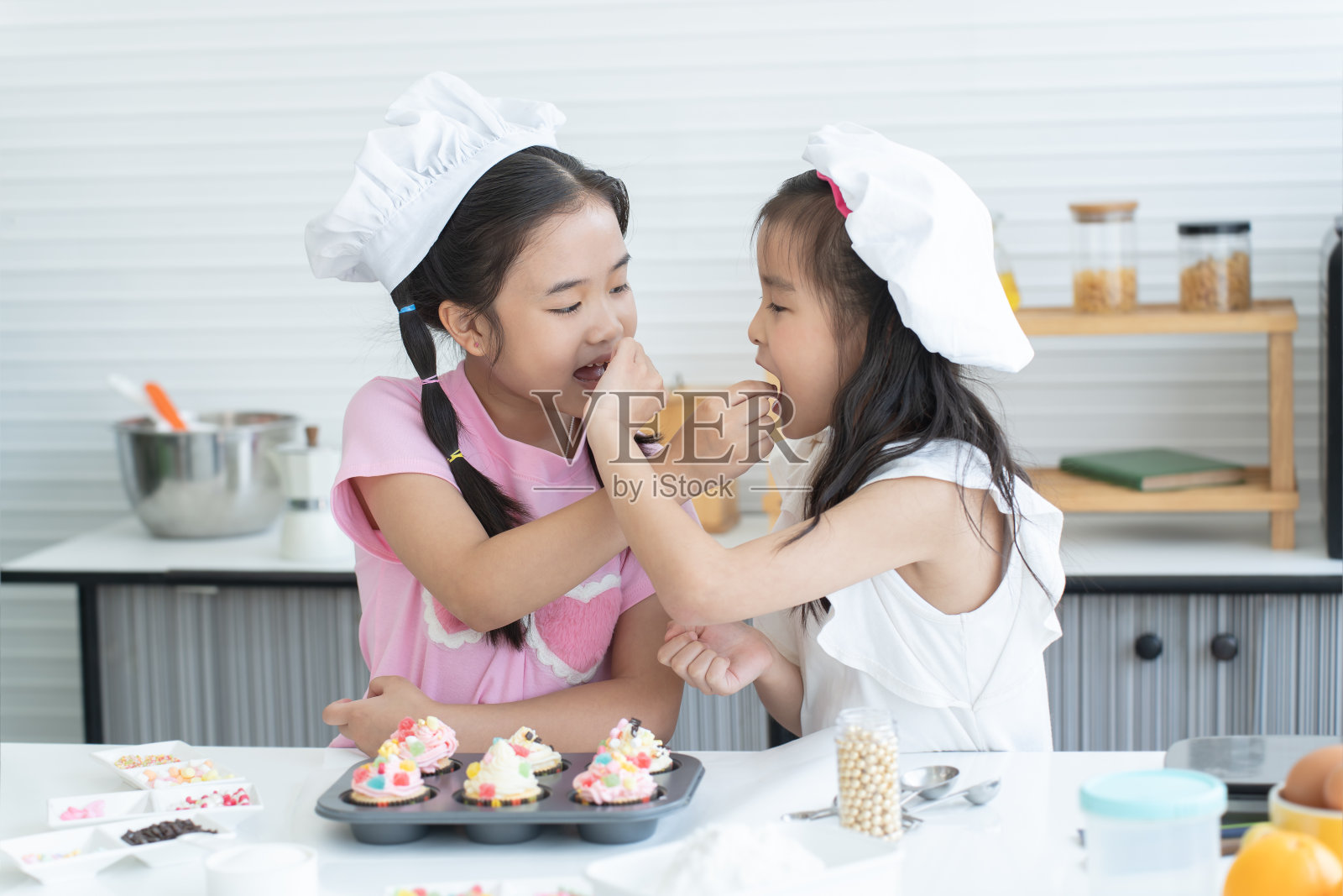 两个可爱的姐妹戴着厨师帽，一起在厨房里用糖粉装饰蛋糕。小女孩们互相喂果冻玩得很开心。快乐的亚洲家庭照片摄影图片