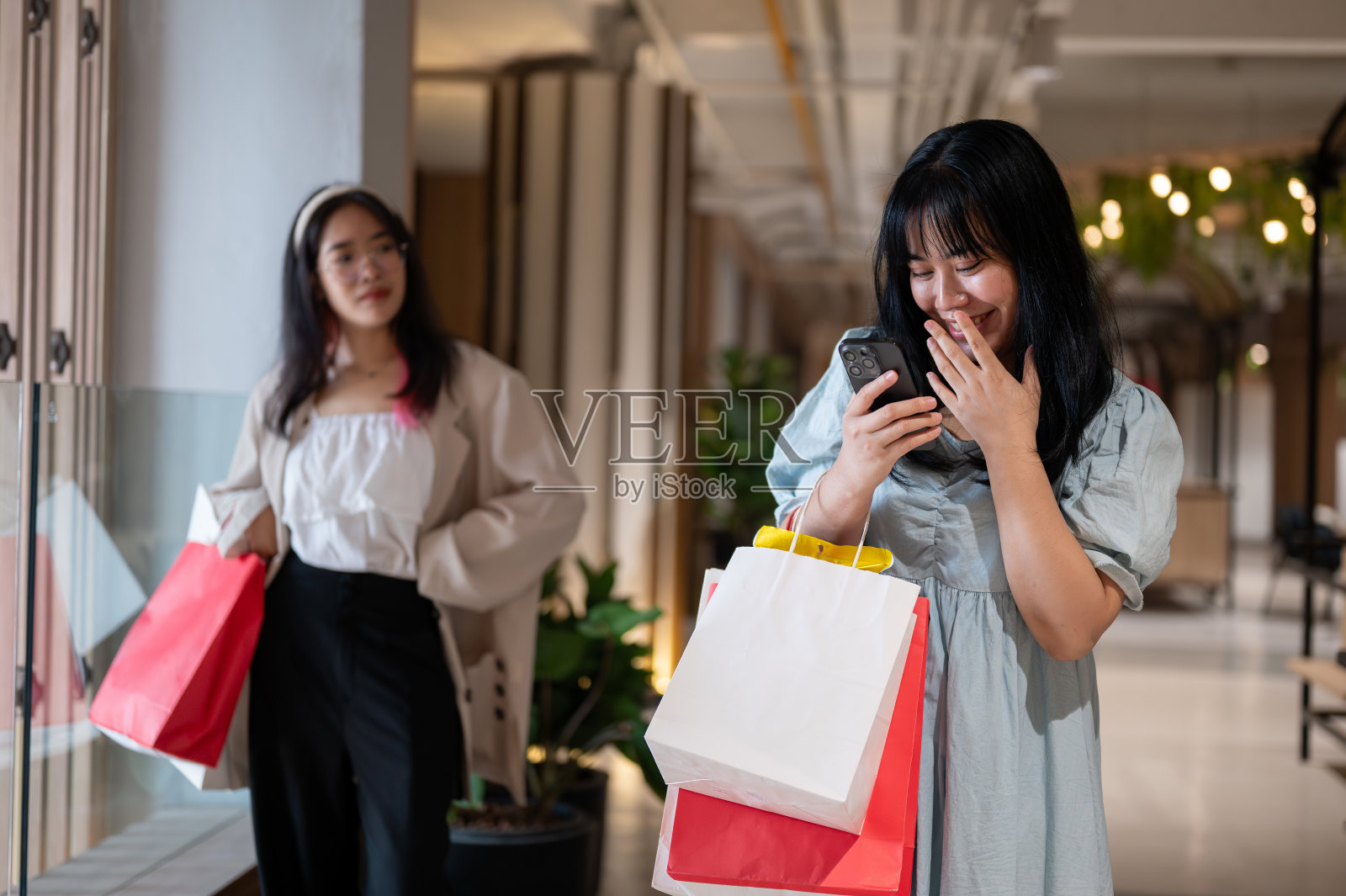 年轻女子看着她的手机，微笑着用手捂着嘴，手里拿着购物袋照片摄影图片