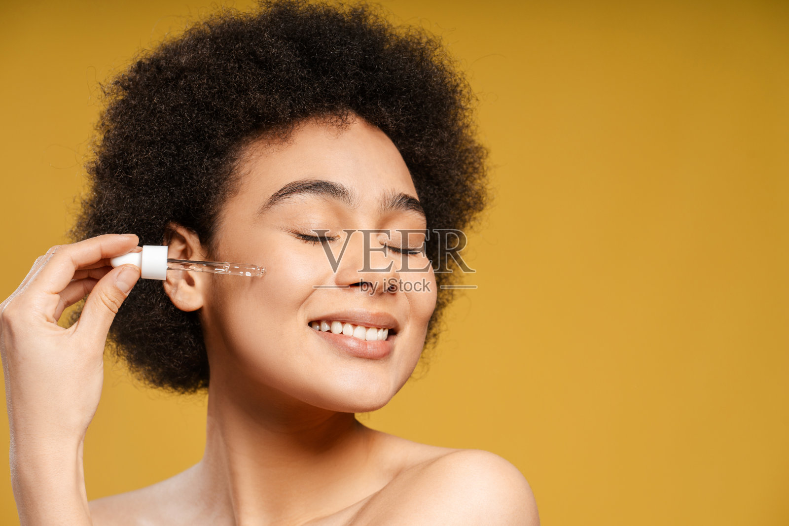 拉丁卷曲头发的年轻女子拿着绿色的瓶子在脸上抹油，穿着白色的毛巾照片摄影图片