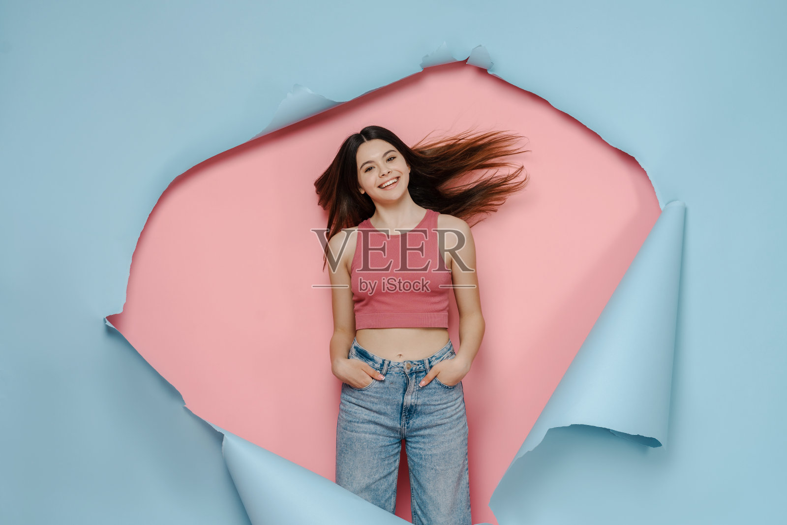 年轻的女孩穿着粉红色的上衣和牛仔裤，双手插在口袋里，在蓝粉色的背景下孤独地看着远方照片摄影图片