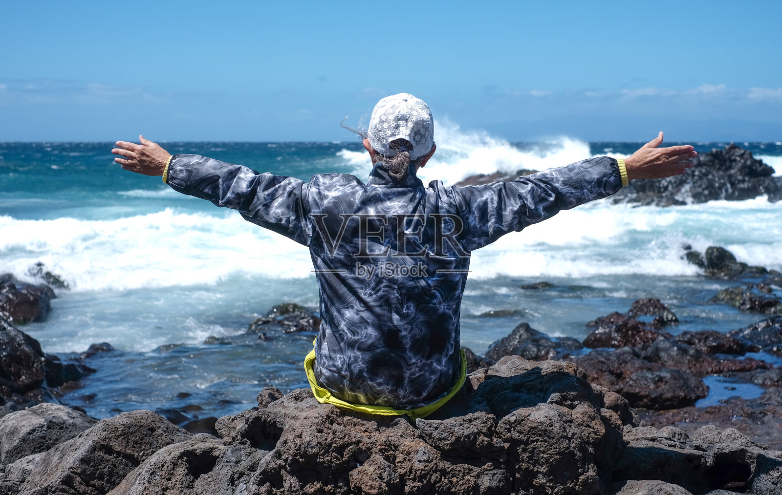 一个成熟的女人伸开双臂坐在岩石沙滩上，欣赏着海浪和大自然的力量。在波涛汹涌的大西洋上，晴朗多风的一天照片摄影图片