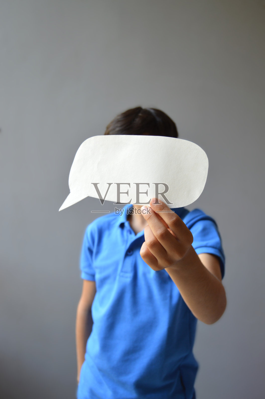 一个穿着休闲蓝色t恤的11岁男孩的垂直照片，他拿着一张剪纸，剪纸的形状是一个空白的、空的演讲泡泡或呼叫气球，作为面具蒙住了他的眼睛。照片摄影图片