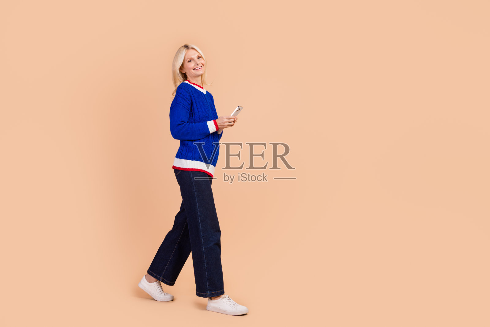 乐观退休人士的全身照片，穿着蓝色套头衫，拿着智能手机，走到米色背景上孤立的空白空间照片摄影图片