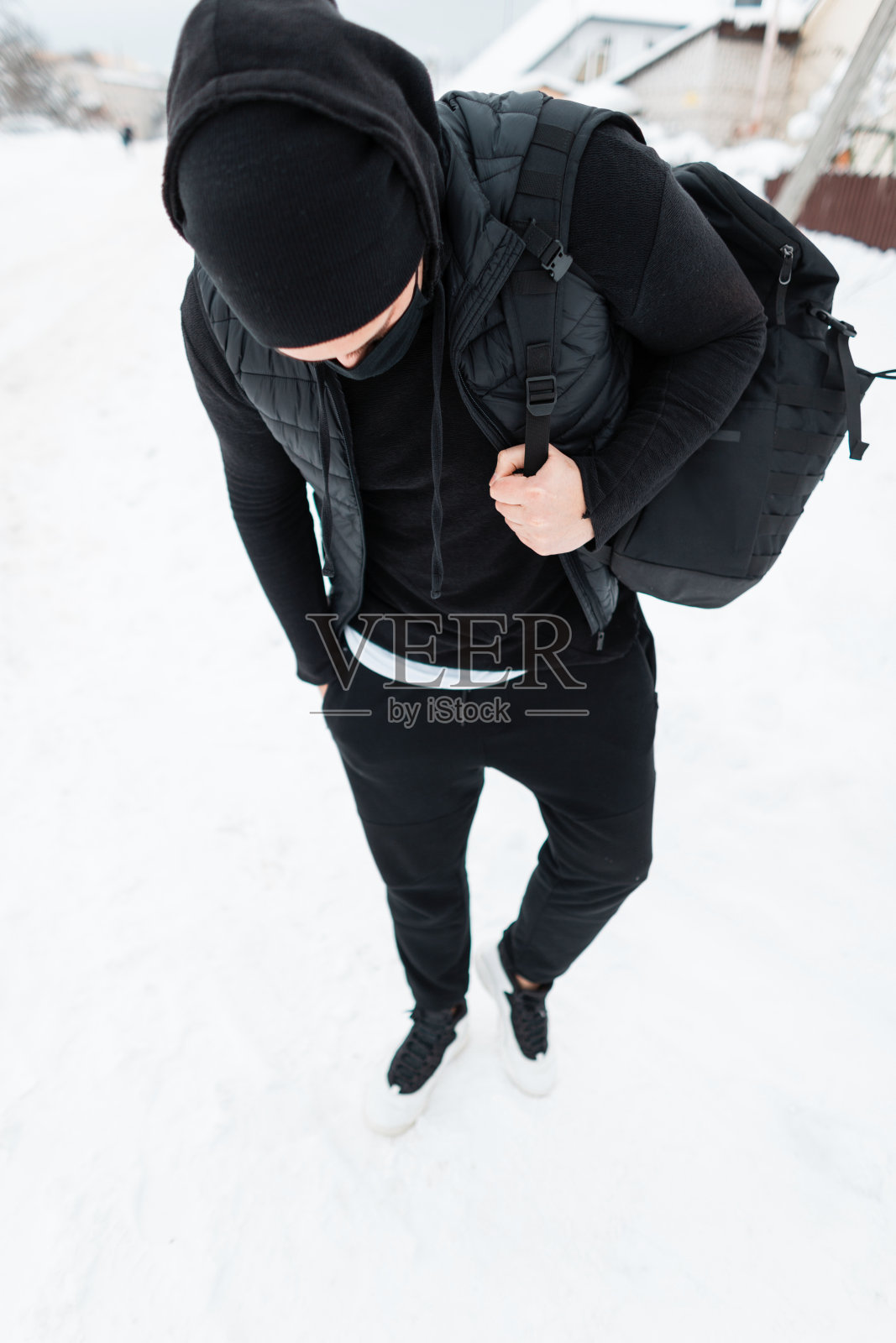 穿着时尚黑色衣服，背着双肩包，戴着防护面具的英俊青年在下雪的冬日里走着。匿名男性隐藏风格照片摄影图片