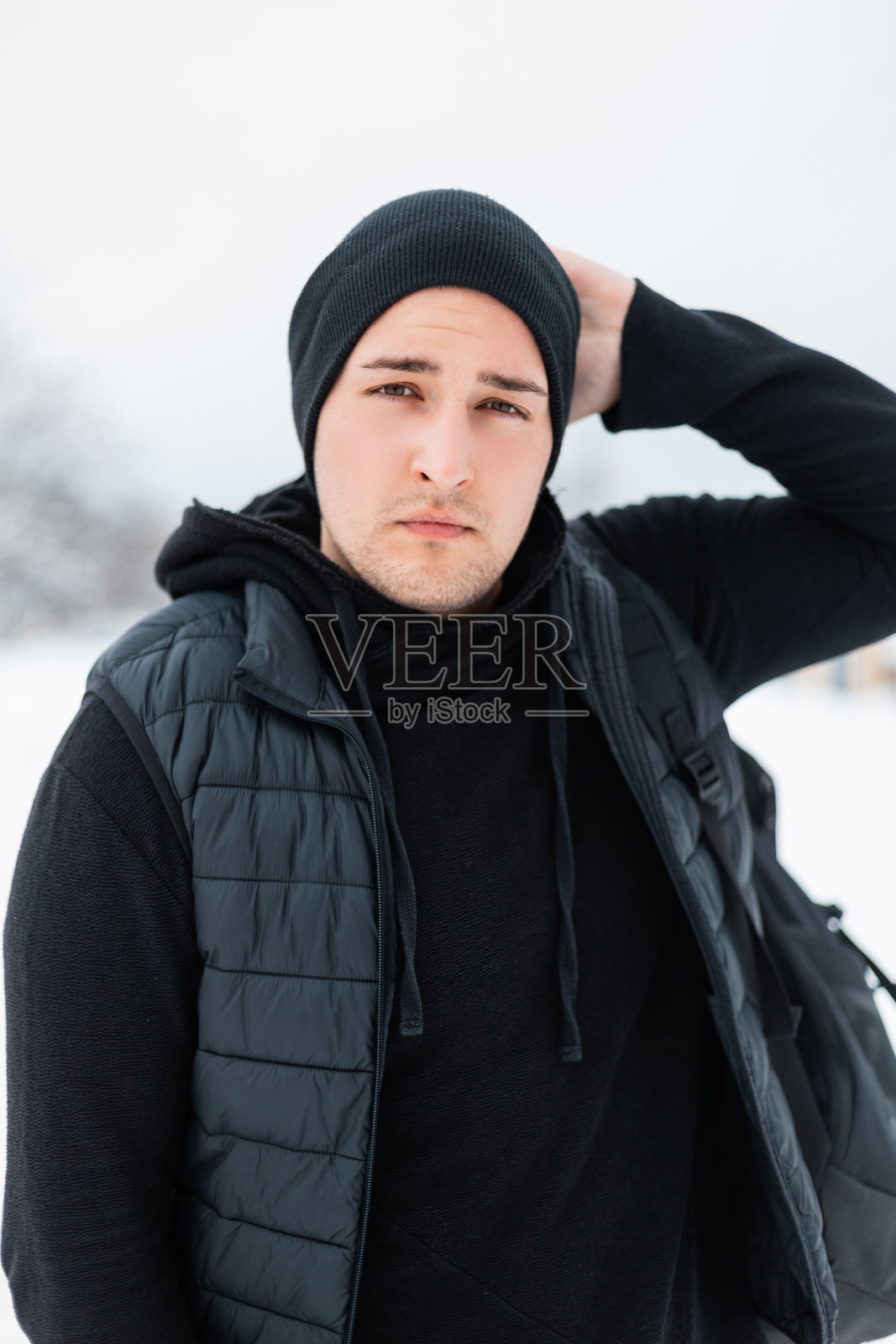 冬日里，穿着黑色冬装的时尚年轻人戴着帽子，穿着卫衣和时尚的背心，背景是白色的白雪照片摄影图片