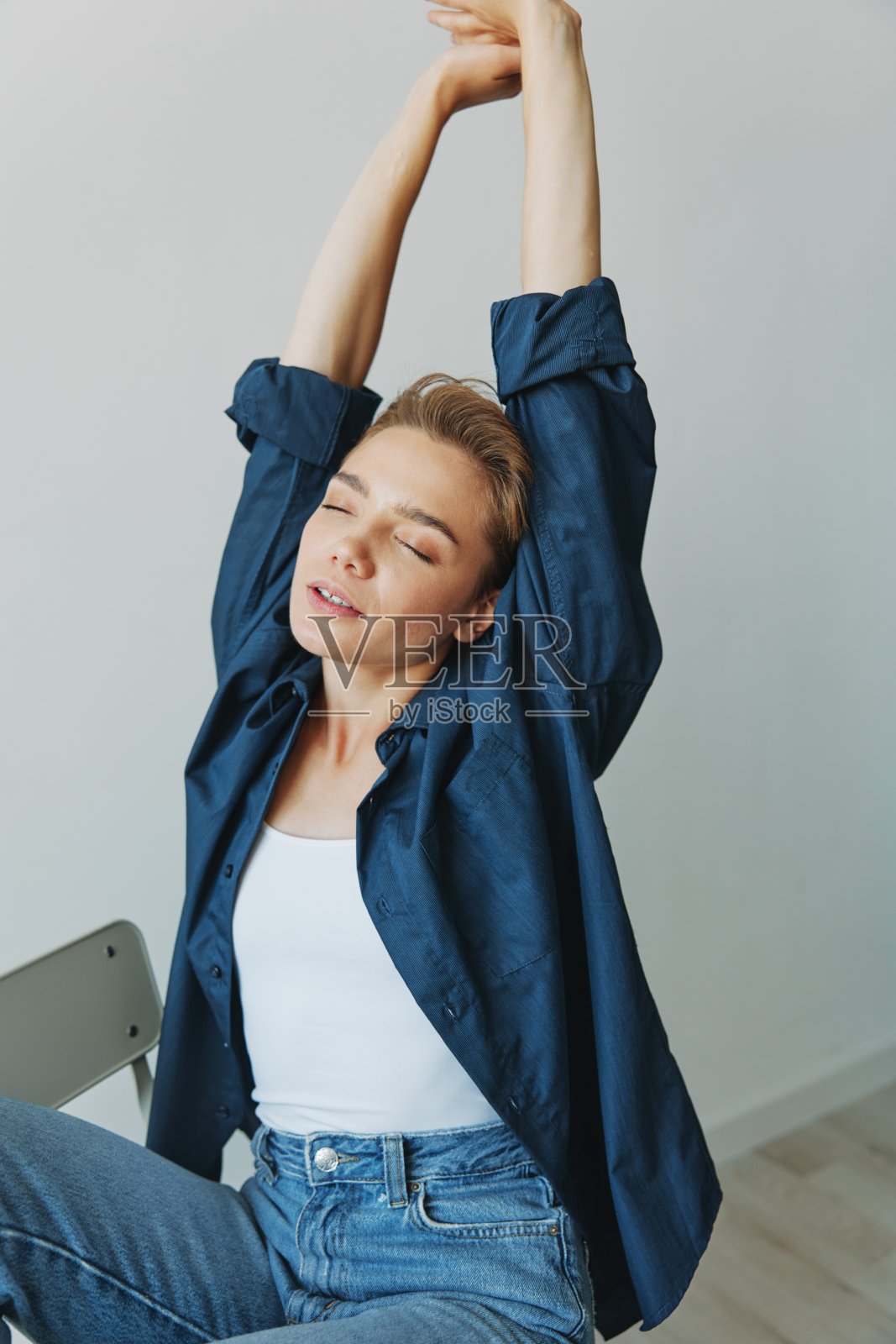 一个年轻的女人坐在家里的椅子上，微笑着，牙齿，短发，穿着牛仔裤和牛仔衬衫，白色背景。没有滤镜的女孩自然姿势照片摄影图片