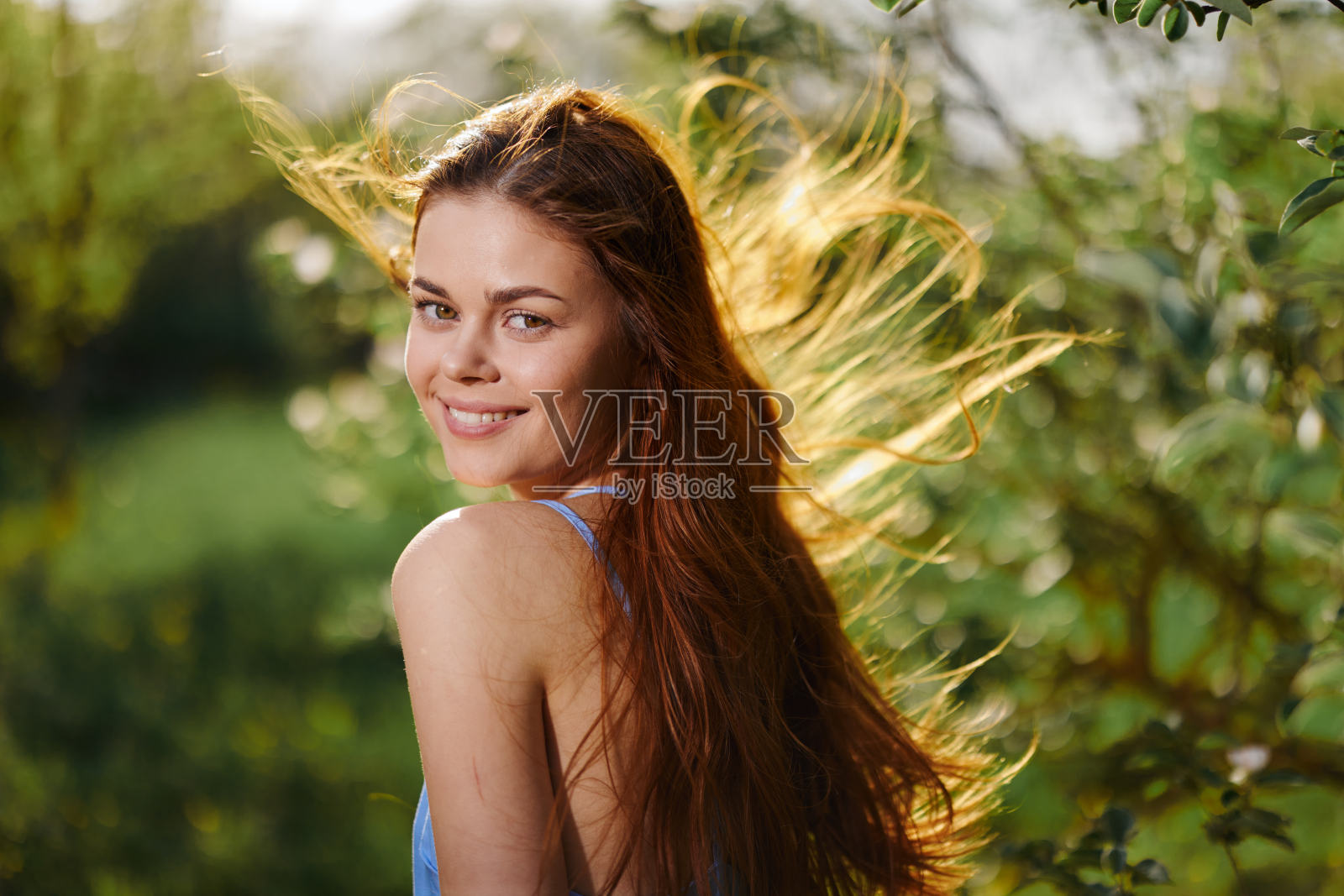 女人带着美丽的微笑，长着牙齿和长发，在春天的夕阳下，在自然公园里，在开花的树旁快乐照片摄影图片