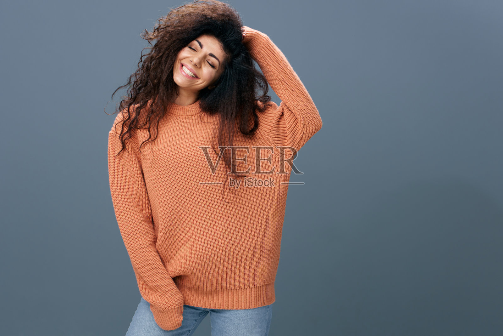 开朗，相当快乐的棕色卷曲的拉丁女性在温暖的毛衣触摸柔软的头发摆出孤立的灰色蓝色背景闭上眼睛。复制空间模拟横幅。时尚为人们提供了情感概念照片摄影图片