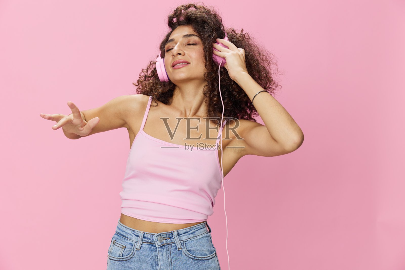 快乐的女人戴着耳机，卷发，闭着眼睛听着音乐和唱歌，穿着粉红色的t恤和牛仔裤，在粉红色的背景DJ派对上，复制空间照片摄影图片