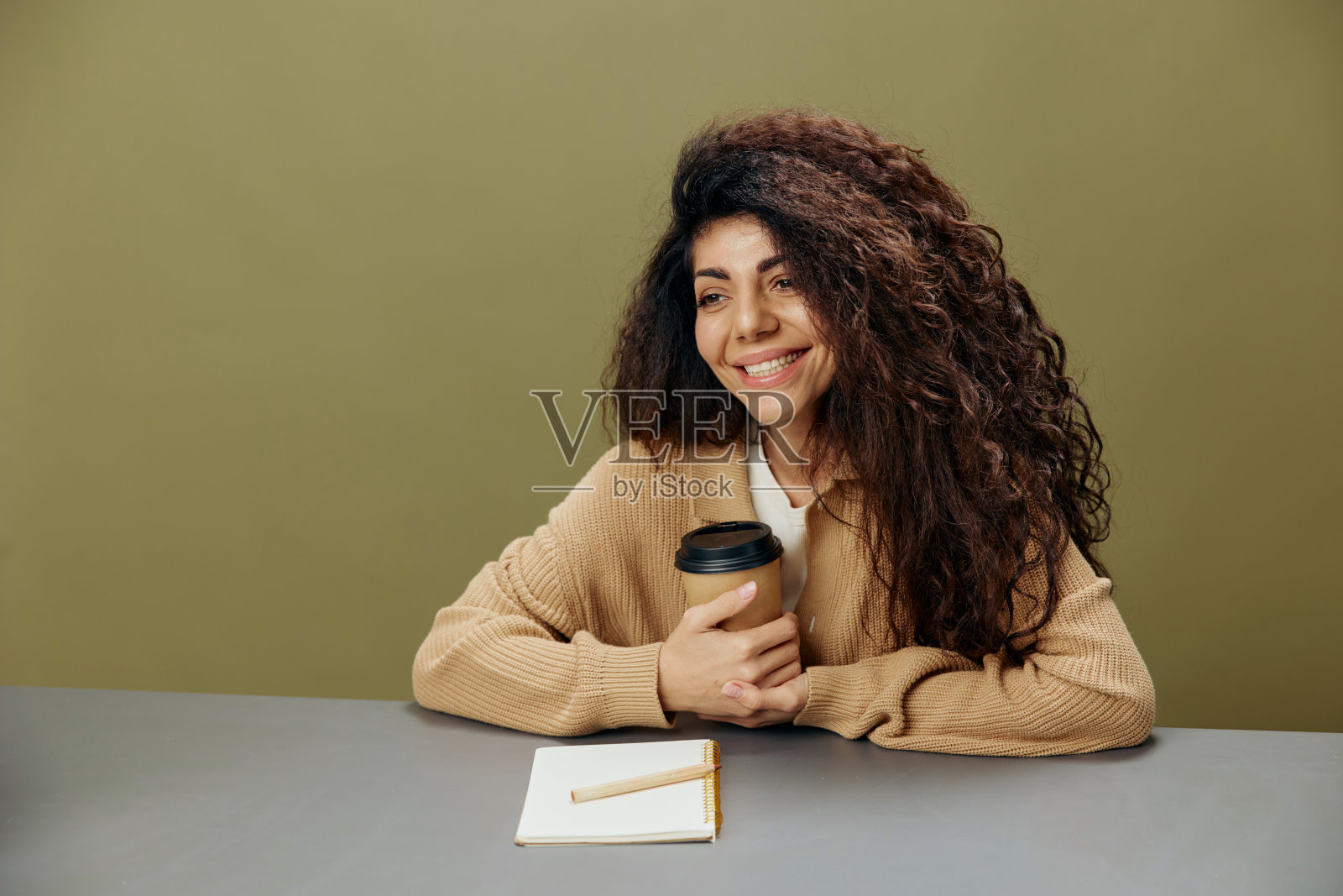 喜欢微笑可爱的古铜色卷发拉丁女士拿着咖啡杯坐在桌子上笔记本孤立在橄榄绿的背景看相机。复制空间模拟横幅。自由在家办公照片摄影图片