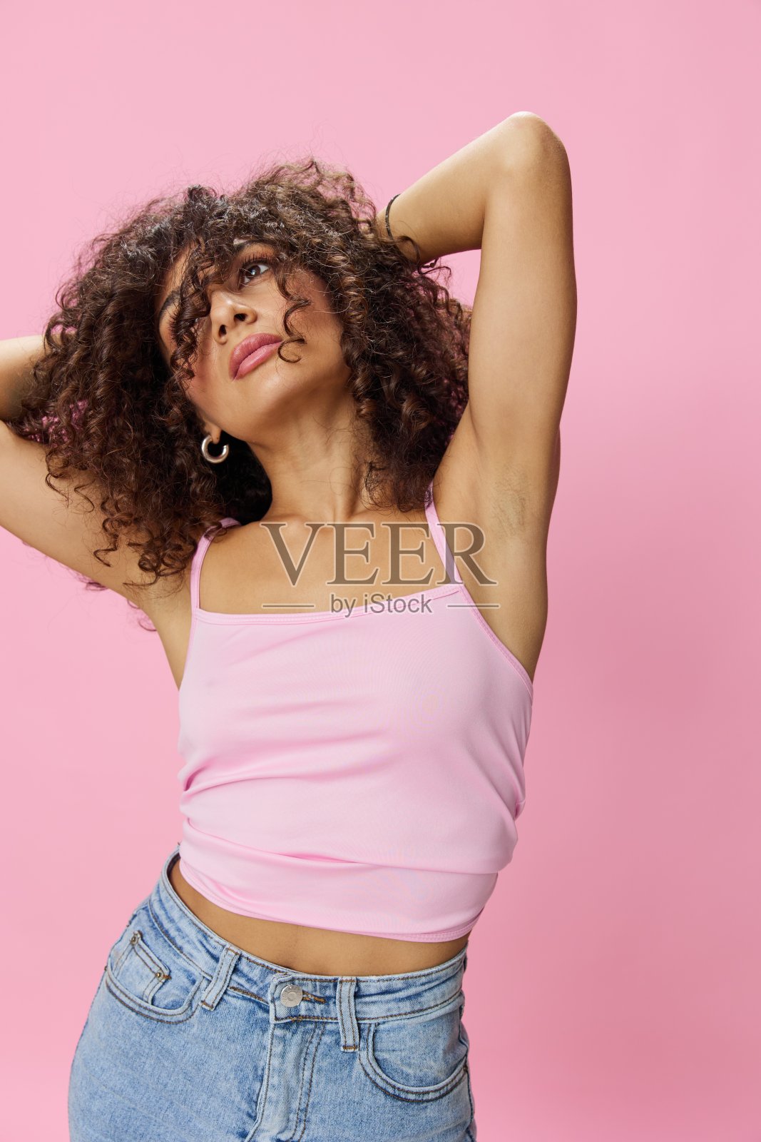 女人卷曲的非洲头发跳舞，粉红色的上衣和牛仔裤在粉红色的背景，微笑，复制的地方照片摄影图片