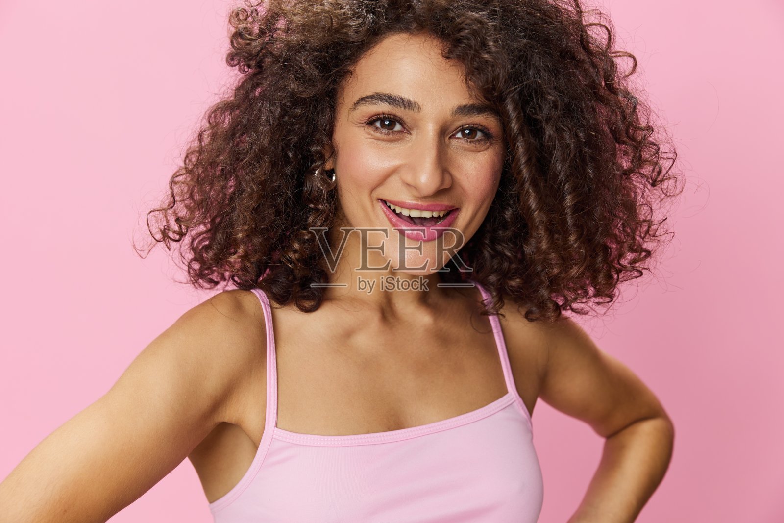 有着卷曲非洲发型的女性模特穿着粉色t恤在粉色背景上摆姿势，自由的动作和舞蹈，看着镜头，微笑着露出牙齿和幸福，复制空间照片摄影图片