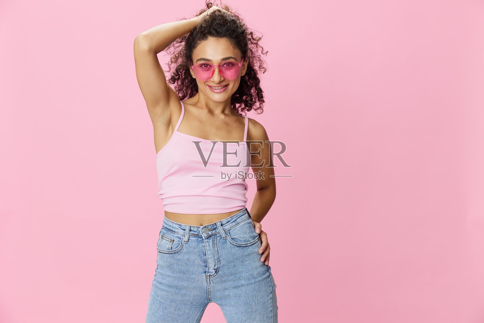 快乐的女人举起手给腋窝打蜡，剃须，卷发，粉色背心和粉色底色牛仔裤，戴着太阳镜，上面有一个漂亮的棕褐色，复制空间照片摄影图片