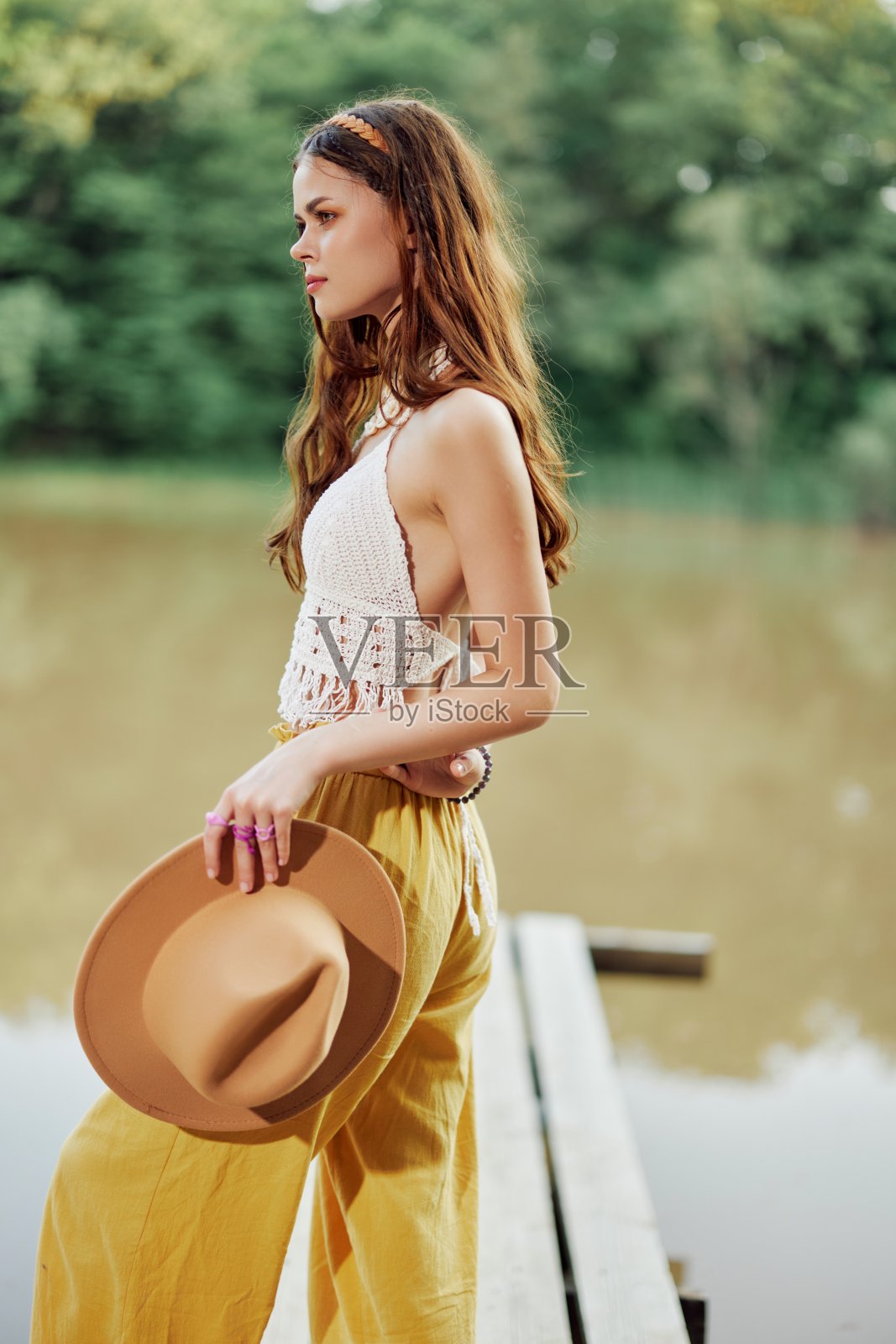 一名年轻女子戴着帽子，穿着黄色的裤子，在夏日的夕阳下，在湖边跳舞，这是一幅嬉皮士的形象，她微笑着照片摄影图片