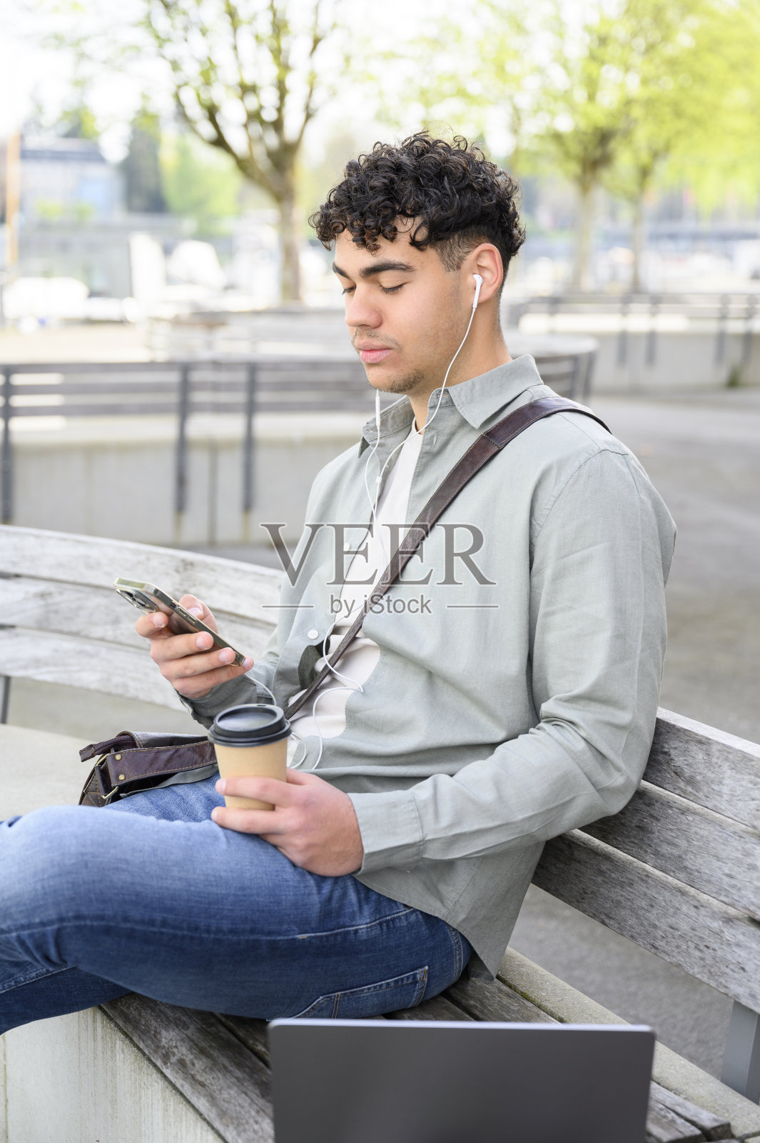 一个年轻人坐在长凳上，手里拿着笔记本电脑和一杯咖啡照片摄影图片