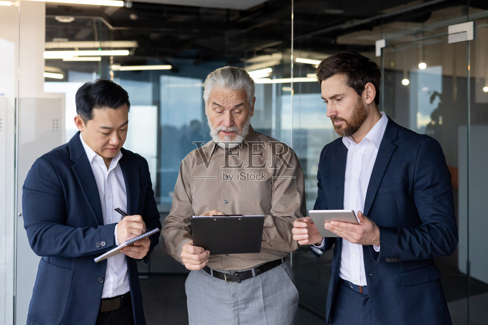 三个不同国籍的年轻商人站在办公室里，手里拿着一个平板电脑和文件，讨论一个联合项目，伙伴关系照片摄影图片