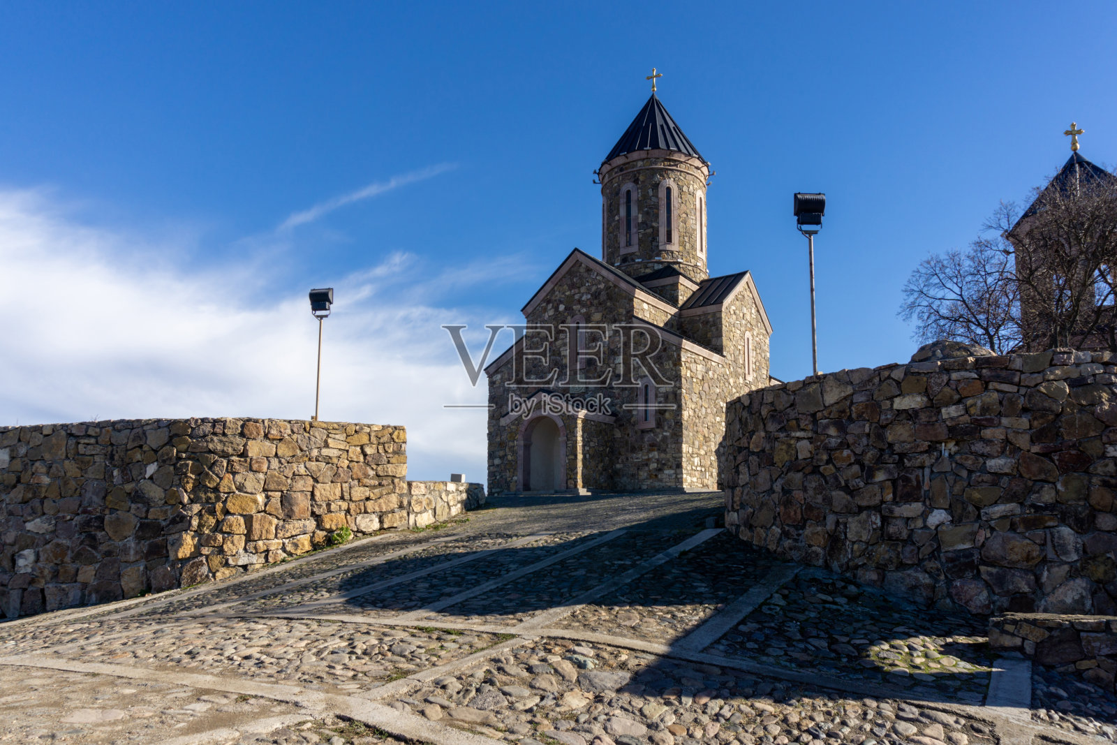 圣彼得和保罗修道院的入口。石铺路，石围栏，背光灯具。明暗相间的石墙，铁穹和镀金的十字架。明亮的一天和蓝色的天空。乔治亚州照片摄影图片