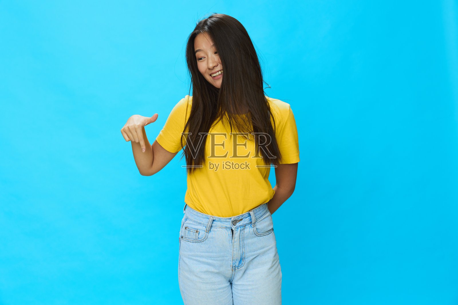 一位身穿黄色t恤和蓝色牛仔裤的亚洲女性指着一个宽松的背景照片摄影图片