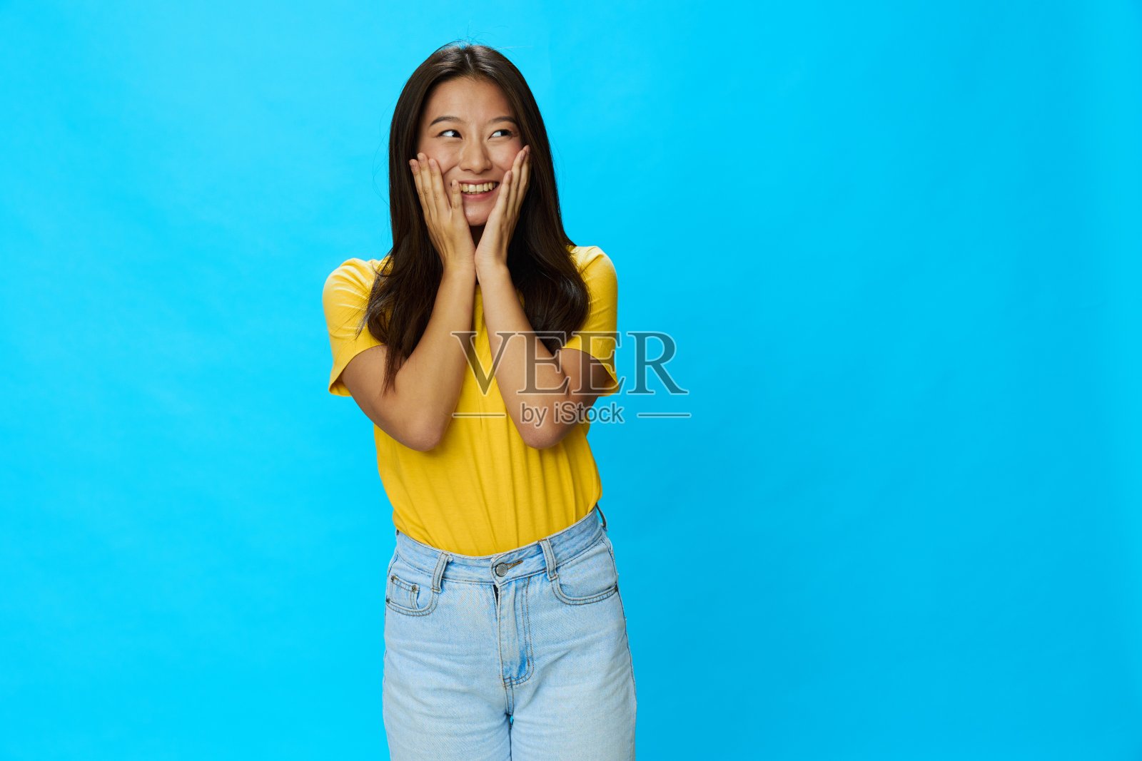 蓝色背景的黄色t恤和蓝色牛仔裤，微笑着露出牙齿的亚洲女人，复制空间照片摄影图片