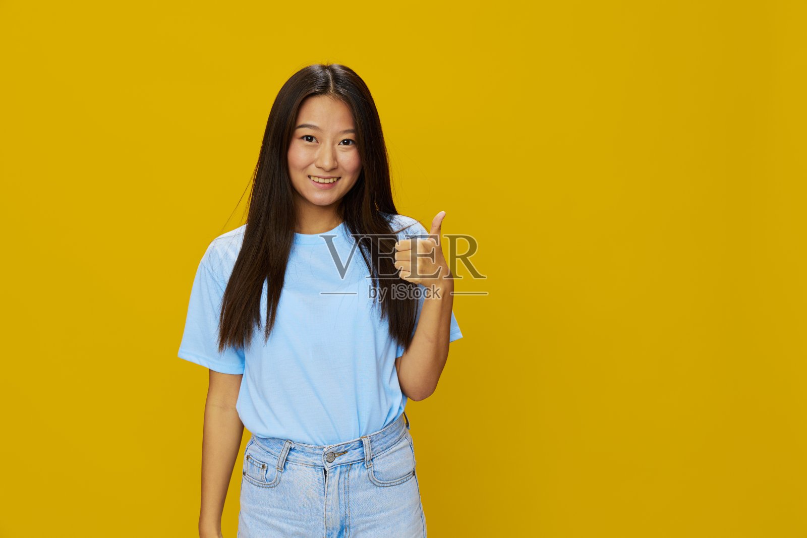 穿着蓝色t恤和牛仔裤的亚洲女性在黄色背景上微笑着，露出牙齿，做手势，做手势和符号照片摄影图片