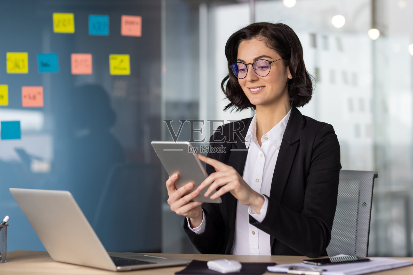 在办公室里穿着西装拿着平板电脑的女商人，在办公室里使用应用程序的女老板，坐在桌子旁拿着笔记本电脑，微笑着工作照片摄影图片