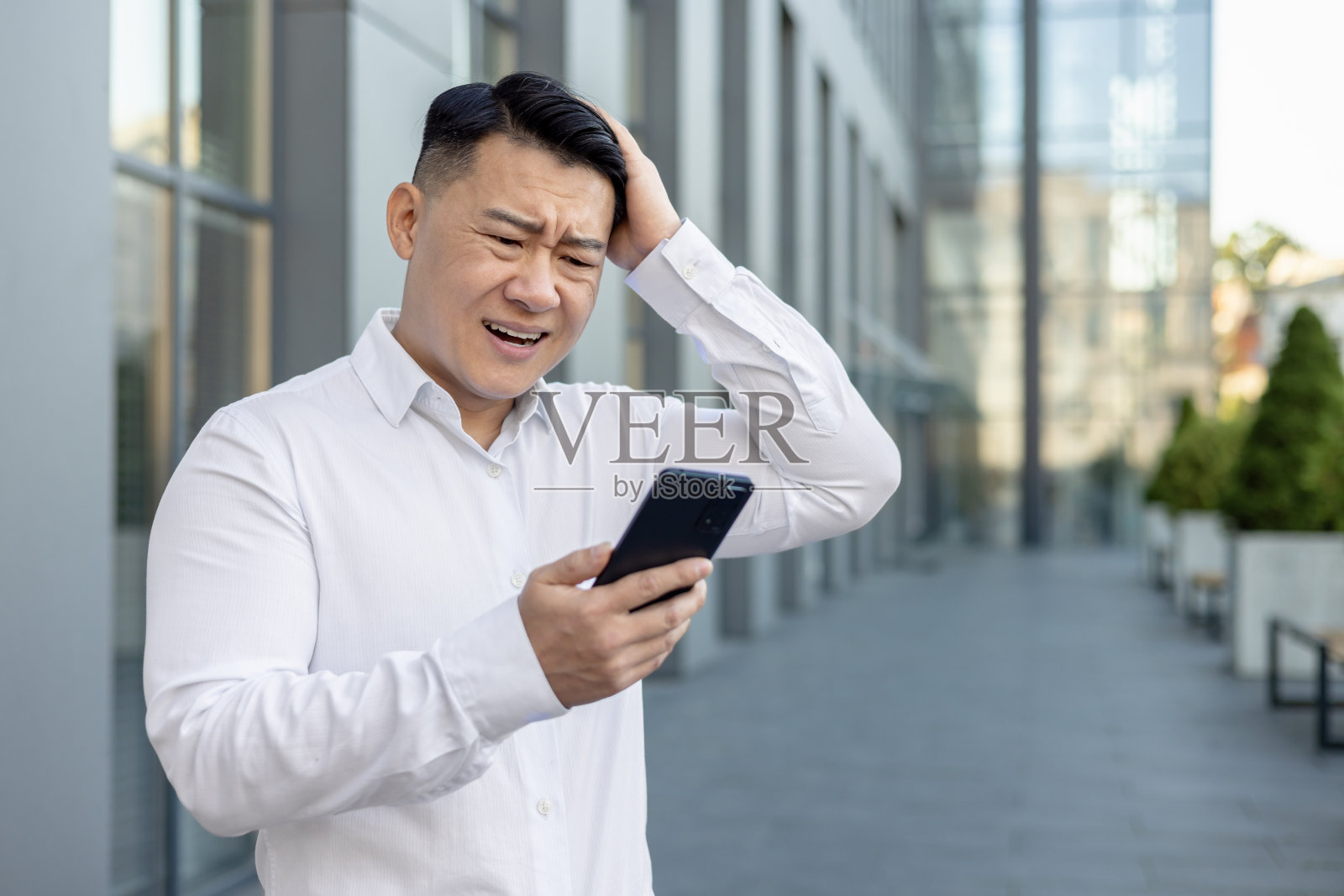 心烦意乱的年轻亚洲人站在办公楼附近，沮丧地看着手机屏幕，忧心忡忡地抱着头照片摄影图片