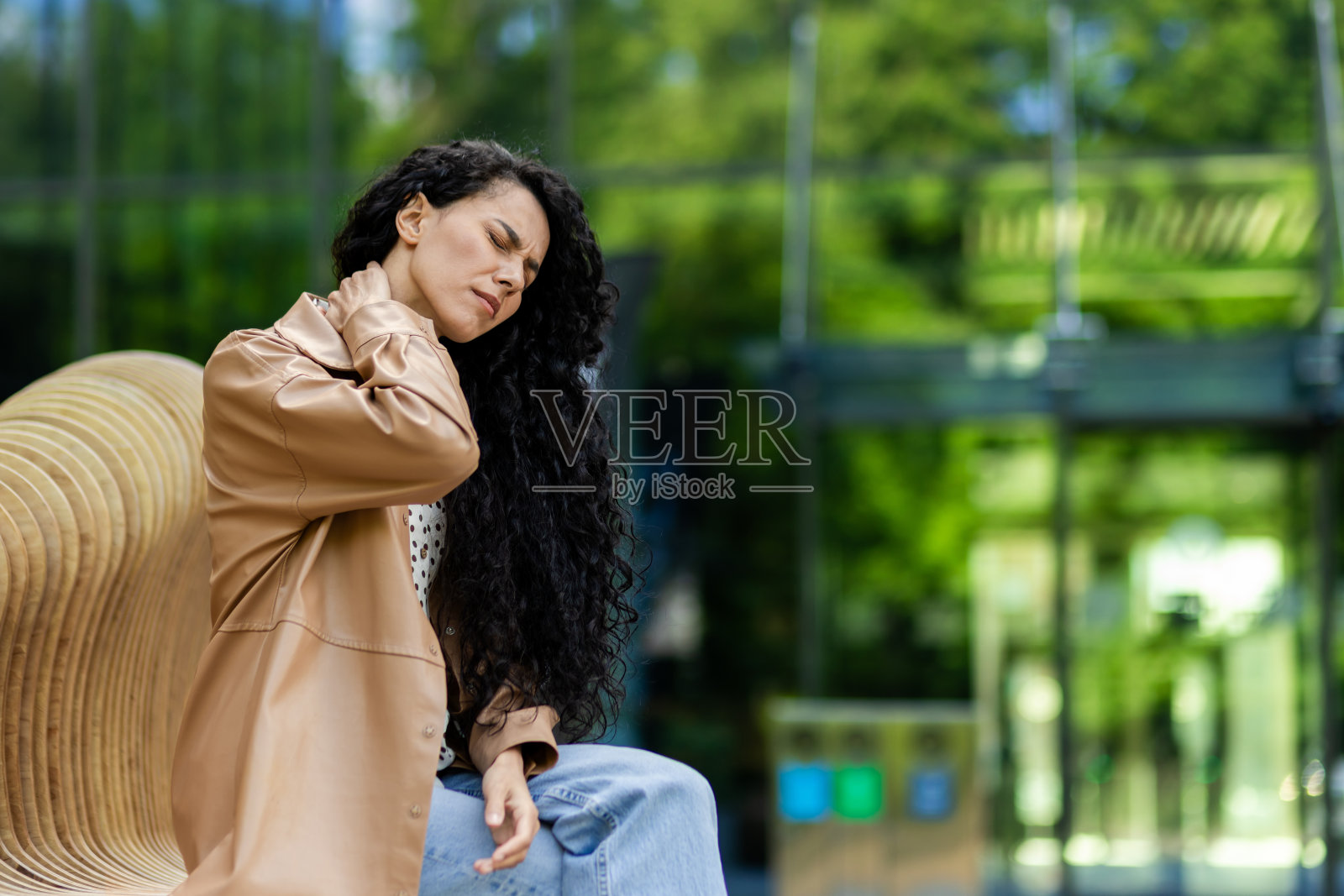 在玻璃建筑附近的木凳上休息时，脖子剧痛的疲惫妇女的侧视图。心烦的女性闭着眼睛在户外按摩疼痛部位照片摄影图片