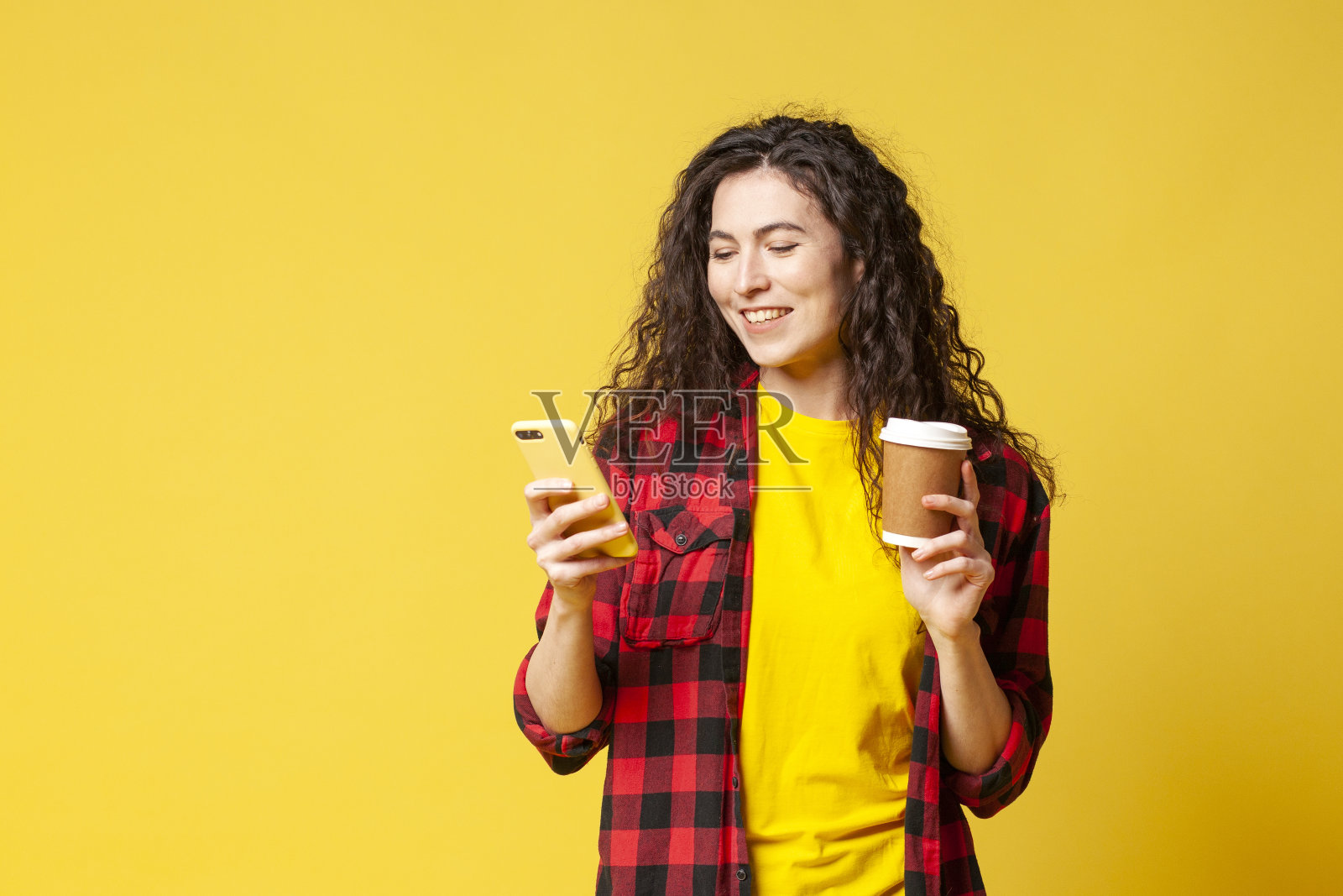 身穿格子衬衫的年轻卷发黑发女孩端着咖啡，用着黄色背景的智能手机照片摄影图片
