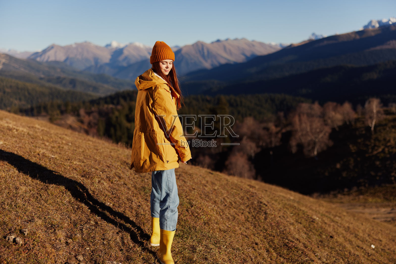 女人笑着带着牙齿全长走幸福走在山上，看山在一个黄色的雨衣和牛仔裤在秋天快乐的旅程在阳光下日落徒步旅行，自由的生活方式照片摄影图片