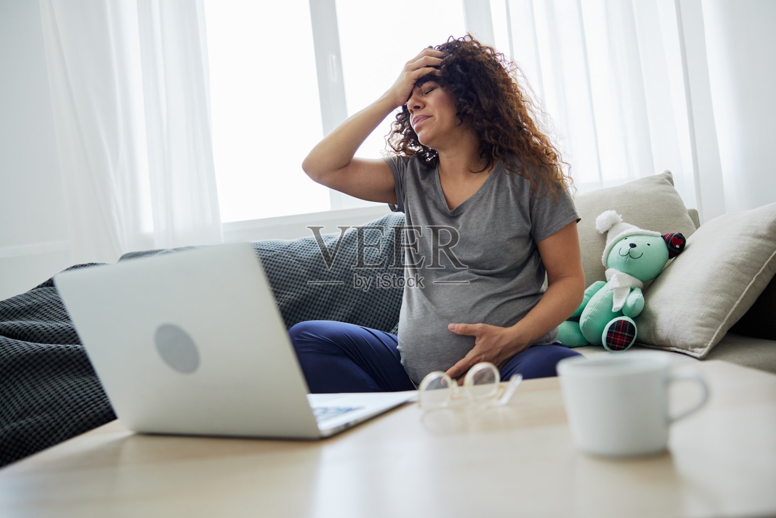 一位头痛的孕妇坐在家里的沙发上，用笔记本电脑看电视节目、电影和连续剧。孕妇的生活方式，准备分娩，怀孕的最后一个月照片摄影图片