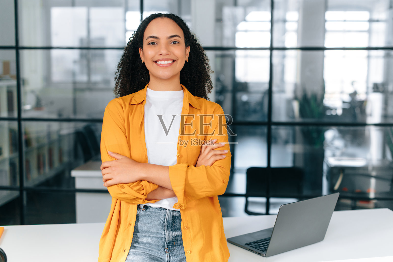 照片上是一位漂亮、积极、自信的巴西或西班牙裔卷发年轻女子，穿着休闲时尚的衣服，站在办公室办公桌附近，双臂交叉，看着镜头，开心地微笑着照片摄影图片