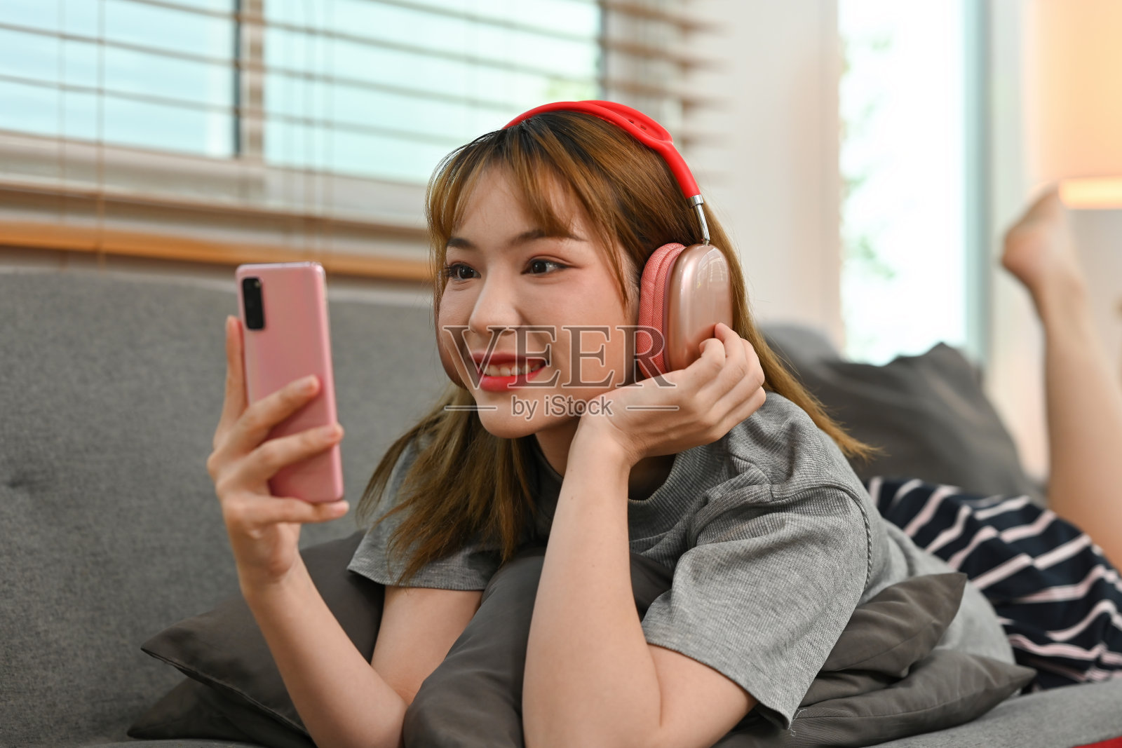 漂亮的少女戴着无线耳机躺在沙发上使用智能手机。照片摄影图片