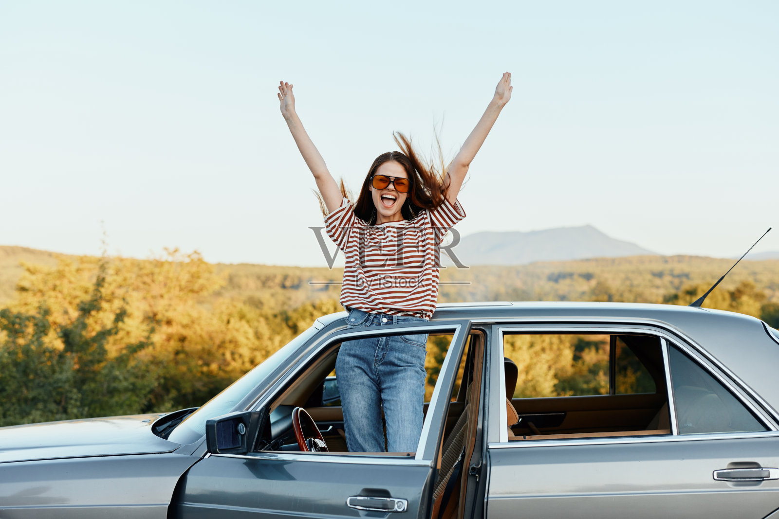 快乐的女旅客爬上汽车，张开双臂开心地笑着。看看周围的大自然。旅行和快乐的生活方式照片摄影图片