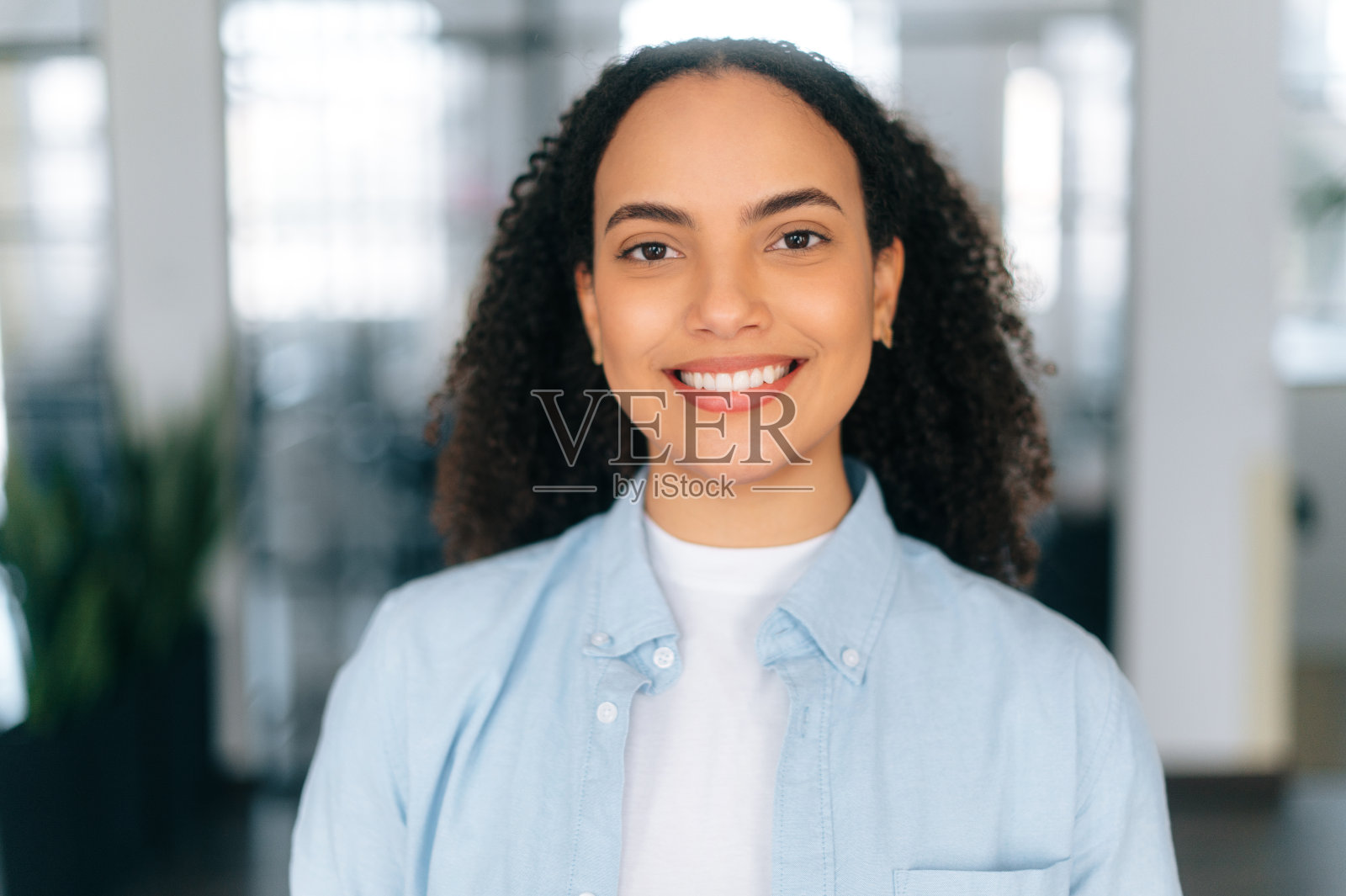 特写照片:漂亮、自信、骄傲的巴西或西班牙卷发女人，穿着淡蓝色衬衫，一家公司的雇员，站在一间现代化的办公室里，看着镜头，友好地微笑着照片摄影图片