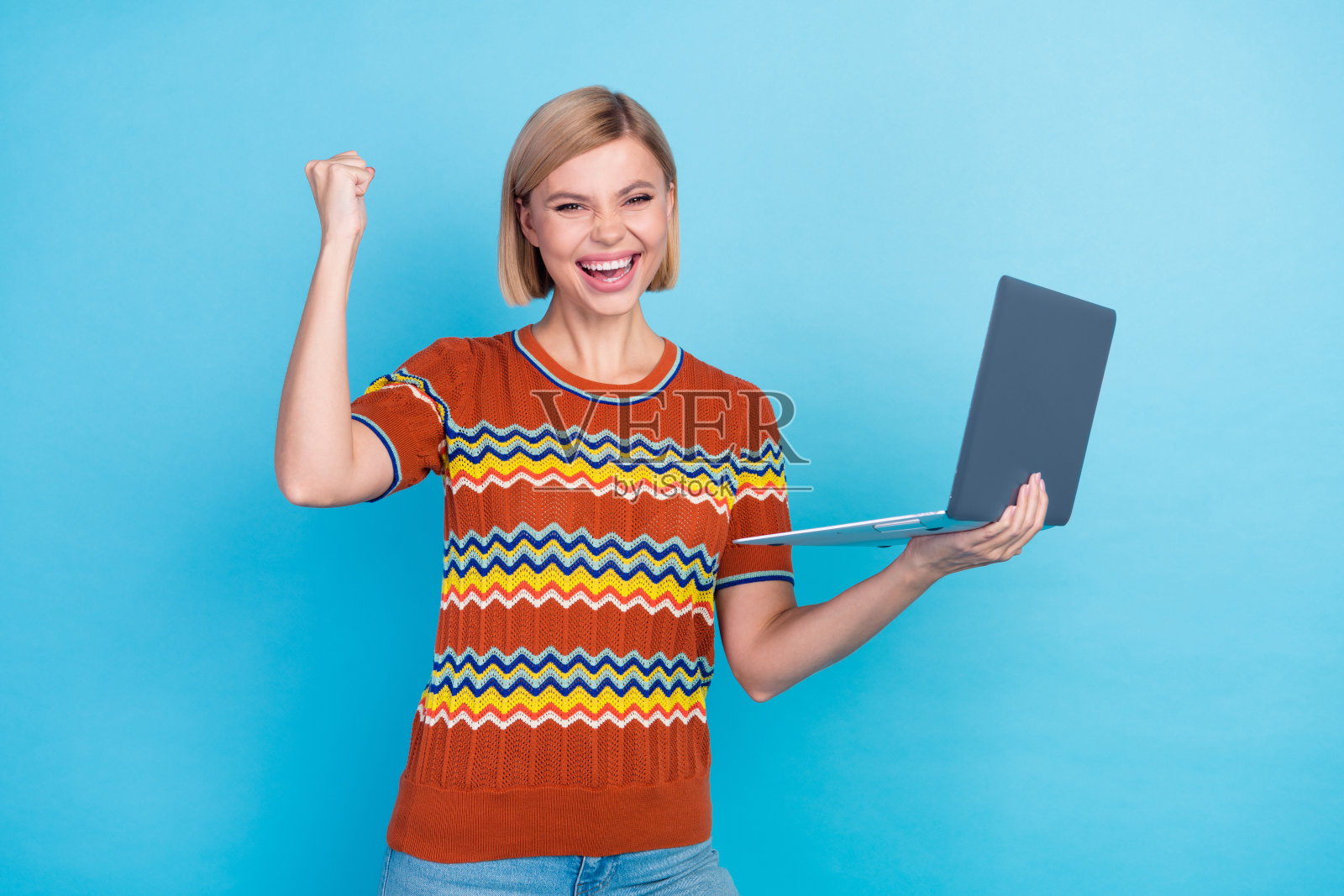 照片中满意的欣喜若狂的女人，梳着鲍勃发型，穿着针织t恤，拿着笔记本电脑，在蓝色的背景上赢得了赌注照片摄影图片